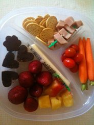 bento breakfast: trawberries and pineapple, chocolate cereal bites, crackers, ham, string cheese, tomatoes and carrots